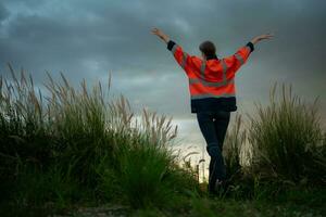 jovem mulher dentro engenheiro uniforme e Alto visibilidade com elevado braços em pé em gramíneo campo às pôr do sol, a conceito do relaxar Tempo depois de trabalhos foto