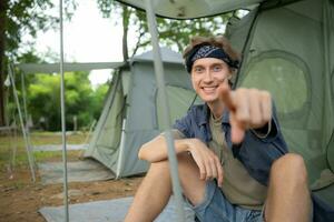 jovem homem sentado dentro frente do barraca às acampamento dentro a campo foto