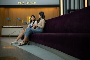 dois jovem ásia mulheres comendo Pipoca e esperando para assistindo filme às frente do caixa escritório foto