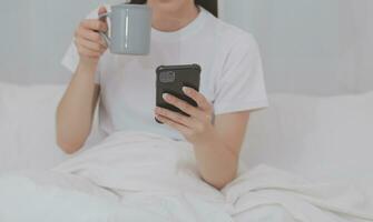 eficiente jovem milenar menina sentado em uma cama dentro a manhã, usa computador portátil computador e come croissants e bebidas café para café da manhã foto