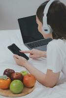 eficiente jovem milenar menina sentado em uma cama dentro a manhã, usa computador portátil computador e come croissants e bebidas café para café da manhã foto