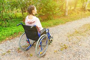 mulher jovem deficiente feliz em cadeira de rodas na estrada no parque hospitalar à espera de serviços ao paciente. menina paralisada na cadeira inválida para pessoas com deficiência ao ar livre na natureza. conceito de reabilitação. foto