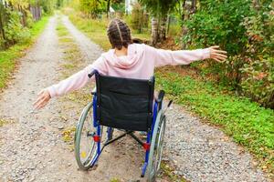 mulher jovem deficiente feliz em cadeira de rodas na estrada no parque hospitalar desfrutando de liberdade. menina paralisada na cadeira inválida para pessoas com deficiência ao ar livre na natureza. conceito de reabilitação. foto