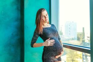 gravidez mulher em pé perto janela às casa e sorrisos foto