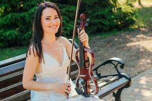 uma mulher artista com Sombrio cabelo dentro uma vestir detém uma de madeira show elétrico violino dentro dela mãos foto