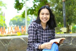 animado jovem ásia mulher Faculdade aluna rindo e sorridente feliz enquanto ler uma livro dentro a parque, Educação conceito foto