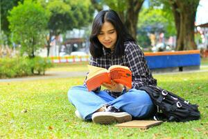 feliz e sucesso fêmea ásia Faculdade aluna desfrutando ler uma livro dentro a parque foto