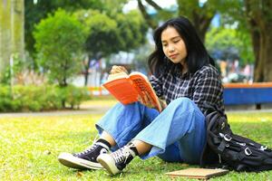 feliz e sucesso fêmea ásia Faculdade aluna desfrutando ler uma livro dentro a parque foto
