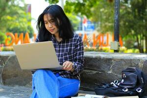 jovem sorridente ásia mulher usando computador portátil e escrevendo caderno, alegre ásia aluna assistindo webinar e estudando a partir de e curso foto