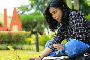 feliz jovem ásia mulher focado usando computador portátil trabalhando remotamente e navegando dentro social meios de comunicação dentro confortável ao ar livre espaço foto