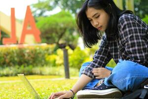 feliz jovem ásia mulher focado usando computador portátil trabalhando remotamente e navegando dentro social meios de comunicação dentro confortável ao ar livre espaço foto