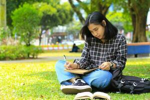 lindo ásia jovem mulher Faculdade aluna focado escrevendo em caderno e lendo livro dentro ao ar livre cidade parque foto