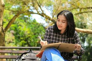 sorridente ásia lindo jovem mulher apreciar escrevendo para Faz Lista e idéia dentro caderno dentro ao ar livre cidade parque foto