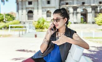 urbano estilo menina sentado em uma Banco chamando em a telefone, a atraente menina sentado falando em a célula telefone, menina sentado dentro uma parque chamando em a telefone foto