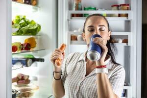 com fome mulher dentro pijamas bebidas Cerveja e come salsichas de a geladeira às noite foto