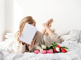 mulher feliz deitada na cama segurando flores de tulipa e um cartão em branco foto