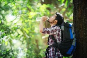 jovem com sede e bebe água durante a caminhada atrás de uma grande árvore. foto