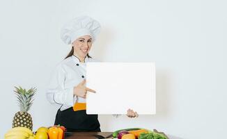 lindo mulher chefe de cozinha com mesa do legumes segurando uma em branco quadro, chefe de cozinha mulher com legumes às mesa segurando em branco cardápio. menina chefe de cozinha dentro a cozinha mostrando uma em branco borda foto