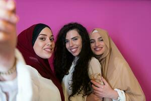 grupo do lindo muçulmano mulheres dois do eles dentro elegante vestir com hijab usando Móvel telefone enquanto levando selfie cenário isolado em Rosa fundo representando moderno islamismo moda tecnologia foto