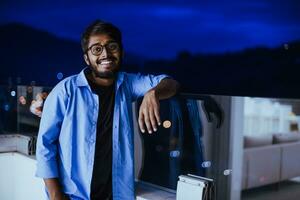 a indiano homem com óculos e uma azul camisa parece por aí a cidade às noite. dentro a fundo do a noite rua do a cidade foto