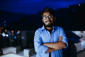 a indiano homem com óculos e uma azul camisa parece por aí a cidade às noite. dentro a fundo do a noite rua do a cidade foto