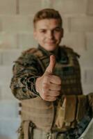 a soldado faz uma gesto do sucesso com dele mão. uma soldado dentro cheio guerra engrenagem carrinhos dentro frente do uma pedra parede e mostra a Está bem placa com dele dedo foto