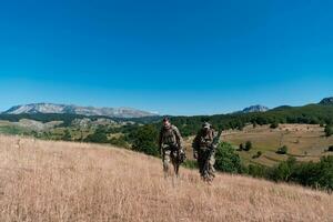 uma Franco atirador equipe pelotão do soldados é indo disfarçado. Franco atirador assistente e equipe líder caminhando e visando dentro natureza com amarelo Relva e azul céu. tático camuflar uniforme. foto