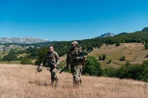 uma Franco atirador equipe pelotão do soldados é indo disfarçado. Franco atirador assistente e equipe líder caminhando e visando dentro natureza com amarelo Relva e azul céu. tático camuflar uniforme. foto