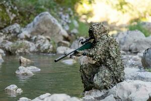 uma militares homem ou airsoft jogador dentro uma camuflar terno esgueirar-se a rio e mira a partir de uma Franco atirador rifle para a lado ou para alvo. foto