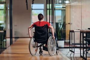 uma moderno jovem empresária dentro uma cadeira de rodas é cercado de a inclusivo área de trabalho com com paredes de vidro escritórios, incorporando determinação e inovação dentro a o negócio mundo foto