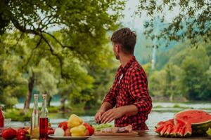 uma homem preparando uma delicioso jantar para dele amigos quem estão tendo Diversão de a rio dentro natureza foto