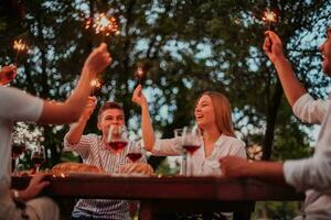 grupo do feliz amigos a comemorar feriado período de férias usando sprinklers e bebendo vermelho vinho enquanto tendo piquenique francês jantar festa ao ar livre perto a rio em lindo verão tarde dentro natureza foto