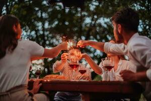 grupo do feliz amigos a comemorar feriado período de férias usando sprinklers e bebendo vermelho vinho enquanto tendo piquenique francês jantar festa ao ar livre perto a rio em lindo verão tarde dentro natureza foto