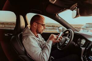 uma homem com uma oculos de sol dirigindo uma carro e tipo uma mensagem em Smartphone às pôr do sol. a conceito do carro viagem foto