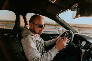uma homem com uma oculos de sol dirigindo uma carro e tipo uma mensagem em Smartphone às pôr do sol. a conceito do carro viagem foto
