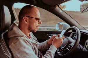 uma homem com uma oculos de sol dirigindo uma carro e tipo uma mensagem em Smartphone às pôr do sol. a conceito do carro viagem foto