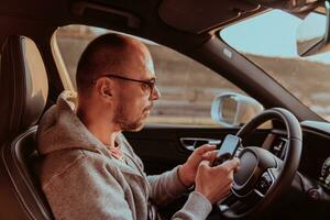 uma homem com uma oculos de sol dirigindo uma carro e tipo uma mensagem em Smartphone às pôr do sol. a conceito do carro viagem foto