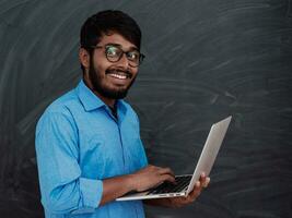 indiano sorridente jovem aluna dentro azul camisa e óculos usando computador portátil e posando em escola quadro-negro fundo foto