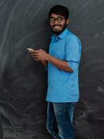 indiano sorridente jovem aluna dentro azul camisa e óculos usando Smartphone e posando em escola quadro-negro fundo foto