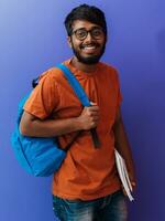 indiano aluna com azul mochila, óculos e caderno posando em roxa fundo. a conceito do Educação e escolaridade. Tempo para ir costas para escola foto