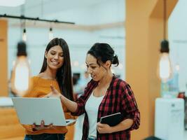 retrato do dois mulheres de negócios falando para cada de outros enquanto em pé dentro uma moderno o negócio escritório com seus colegas usando computador portátil e tábua.. marketing conceito. multiétnico sociedade. foto