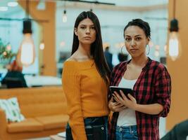 retrato do dois mulheres de negócios falando para cada de outros enquanto em pé dentro uma moderno o negócio escritório com seus colegas usando computador portátil e tábua.. marketing conceito. multiétnico sociedade. foto