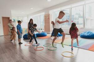 pequeno berçário escola crianças com fêmea professor em chão dentro de casa dentro sala de aula, fazendo exercício. pulando sobre hula aro círculos rastrear em a chão. foto