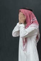 uma jovem árabe homem dentro tradicional roupas fazer uma tradicional oração para Deus mantém dele mãos dentro Rezar gesto dentro frente do uma Preto fundo foto