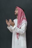 uma jovem árabe homem dentro tradicional roupas fazer uma tradicional oração para Deus mantém dele mãos dentro Rezar gesto dentro frente do uma Preto fundo foto