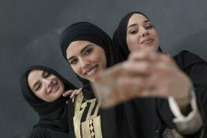 grupo do jovem lindo muçulmano mulheres dentro elegante vestir com hijab usando Smartphone enquanto levando selfie cenário dentro frente do Preto fundo foto