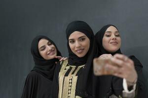 grupo do jovem lindo muçulmano mulheres dentro elegante vestir com hijab usando Smartphone enquanto levando selfie cenário dentro frente do Preto fundo foto