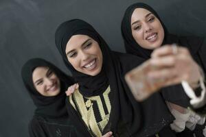 grupo do jovem lindo muçulmano mulheres dentro elegante vestir com hijab usando Smartphone enquanto levando selfie cenário dentro frente do Preto fundo foto