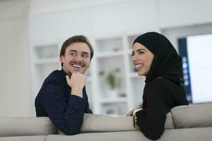 jovem muçulmano casal mulher vestindo islâmico hijab roupas sentado em sofá assistindo televisão juntos durante a mês do Ramadã às moderno casa foto