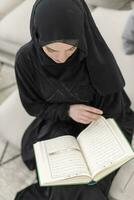 jovem tradicional muçulmano mulher lendo Alcorão em a sofá antes iftar jantar durante uma Ramadã festa às casa foto